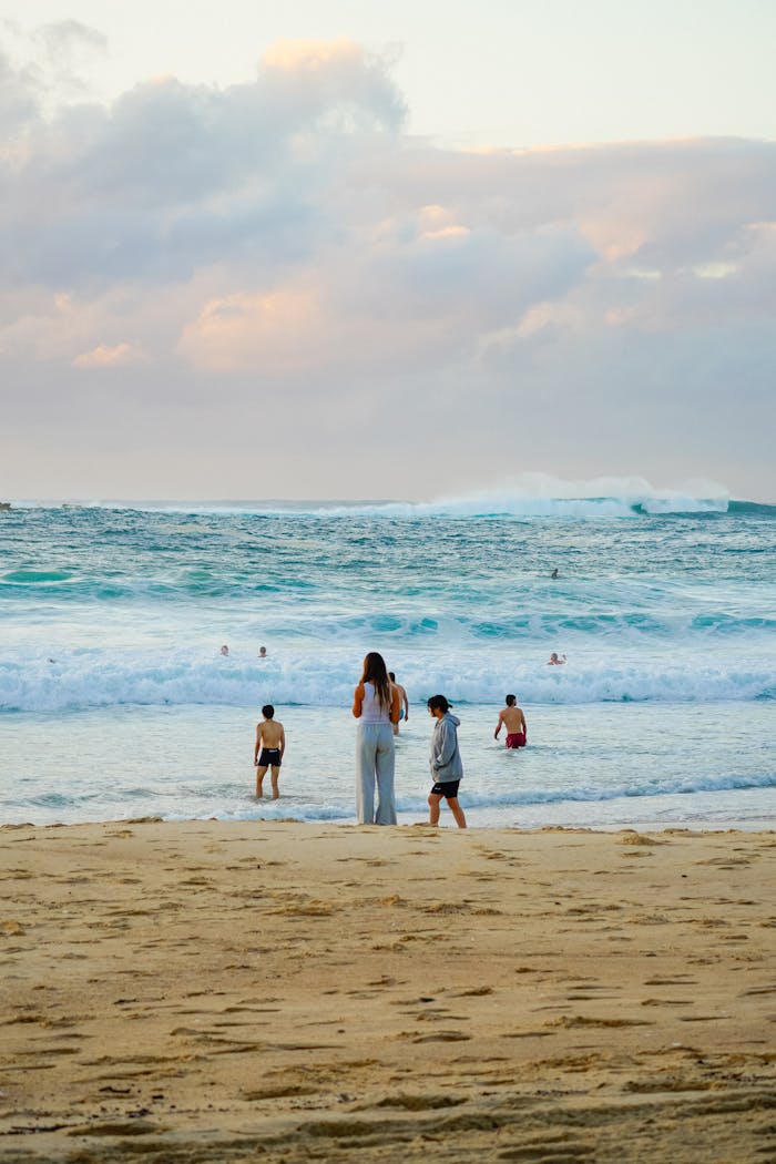 People enjoying a sunny day at the beach with crashing waves and soft clouds.